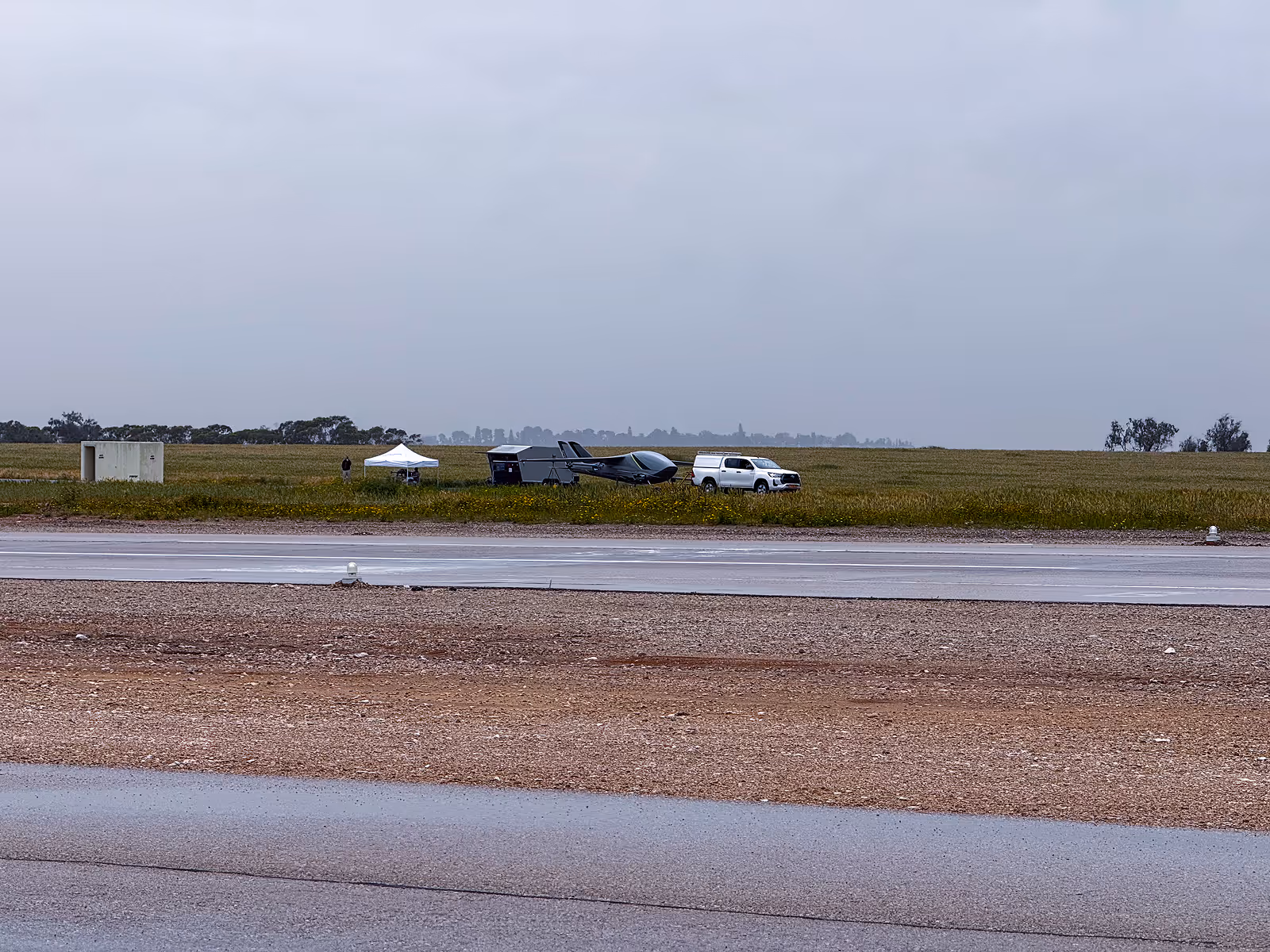 Small gray aircraft and white truck near a white canopy tent in a grassy field by a runway on a cloudy day.
