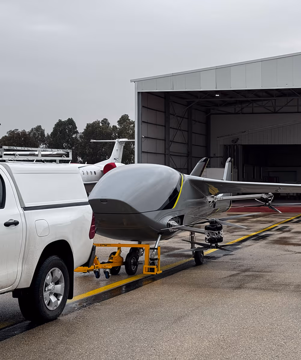 Gray drone aircraft hitched to a white truck near an open aircraft hangar on wet pavement.