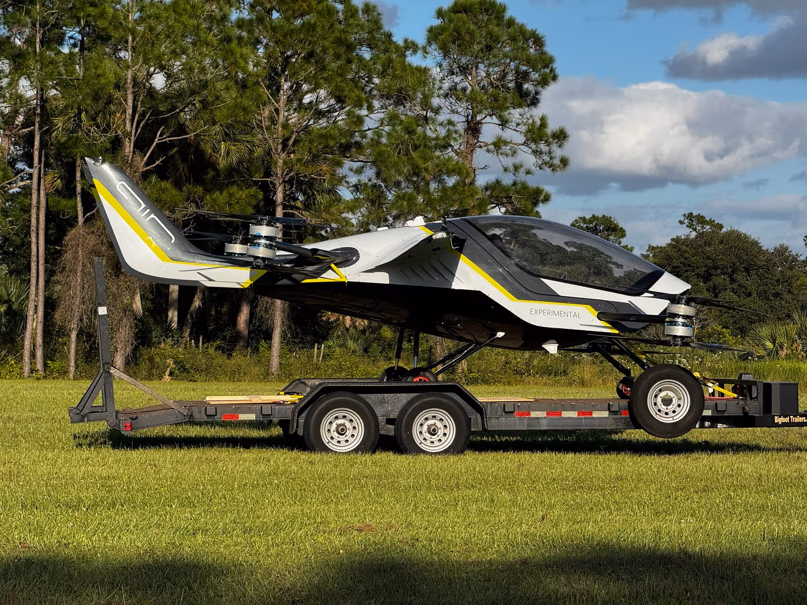 Experimental small aircraft with multiple rotors on a trailer in a grassy area with trees and sky background.