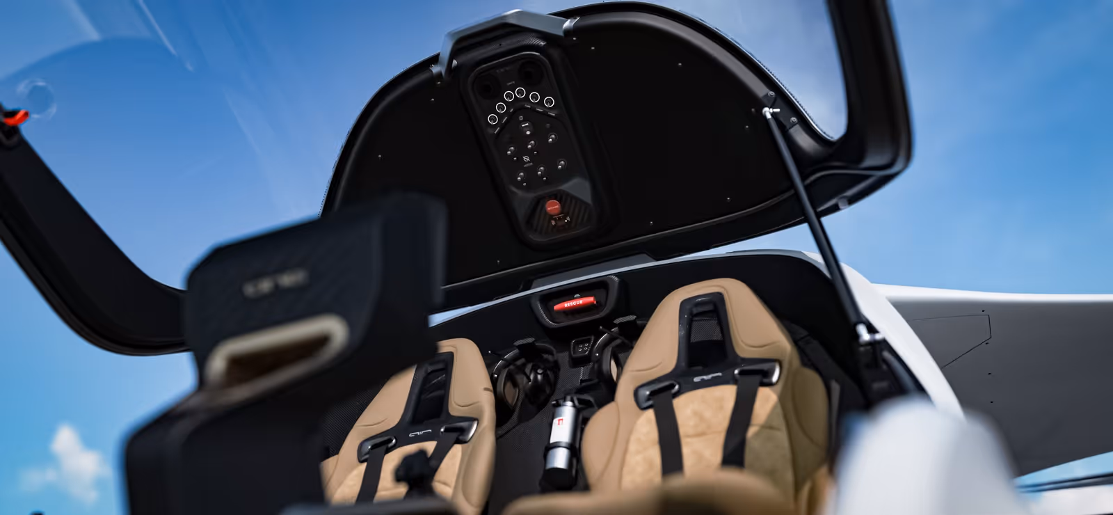 Interior view of a small aircraft cockpit with beige seats and an open canopy against blue sky.