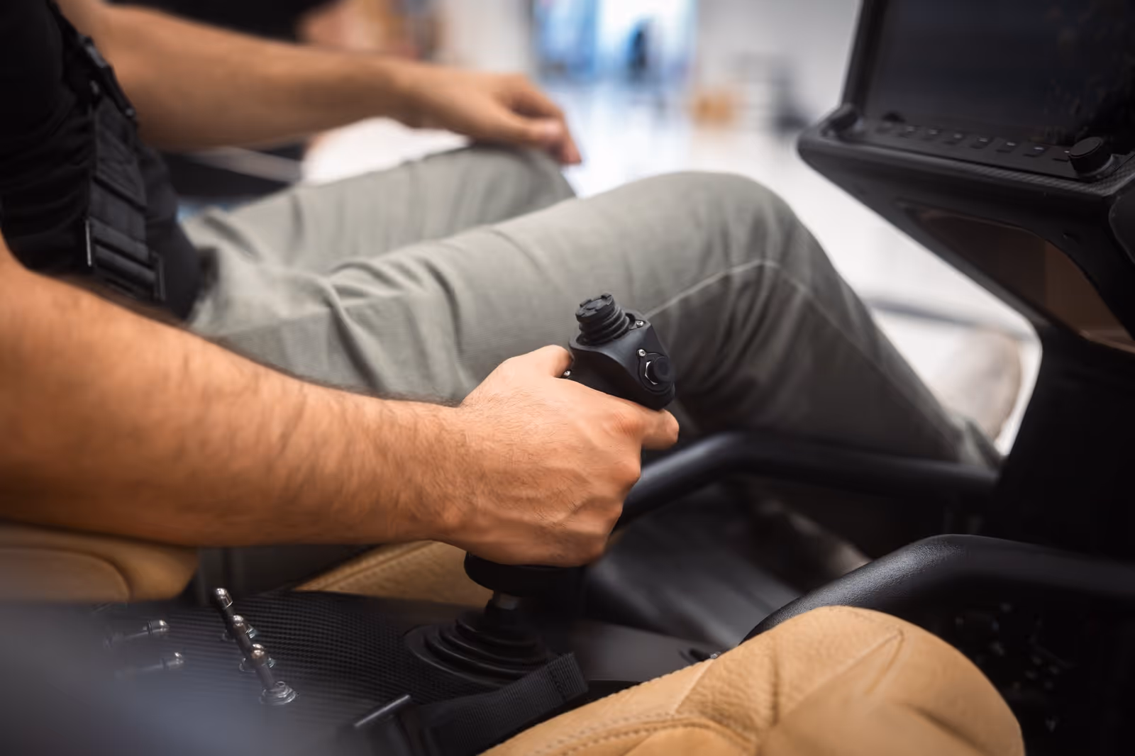 Man's hand gripping joystick inside a vehicle cockpit with tan seats and control switches.