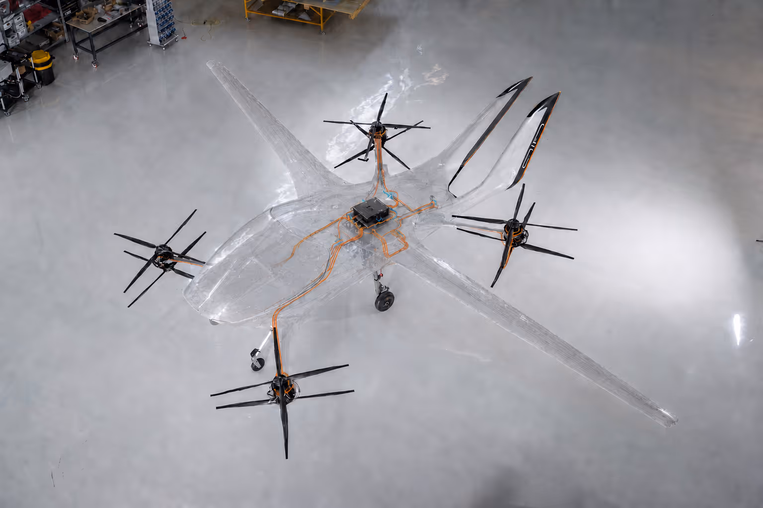Transparent drone with four black propellers and visible wiring on hangar floor.
