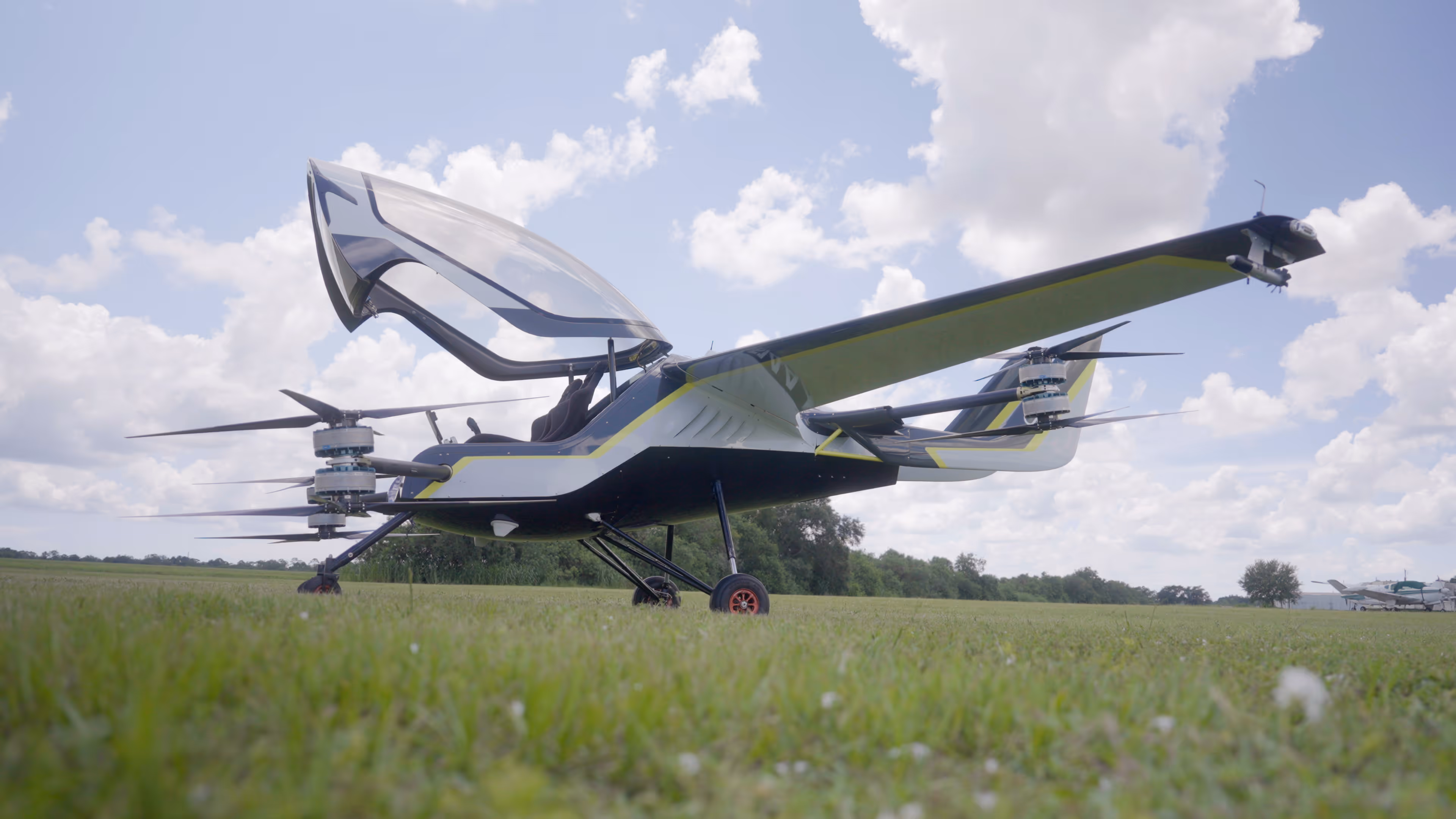 Electric vertical takeoff and landing aircraft on grass under cloudy sky with open cockpit canopy.