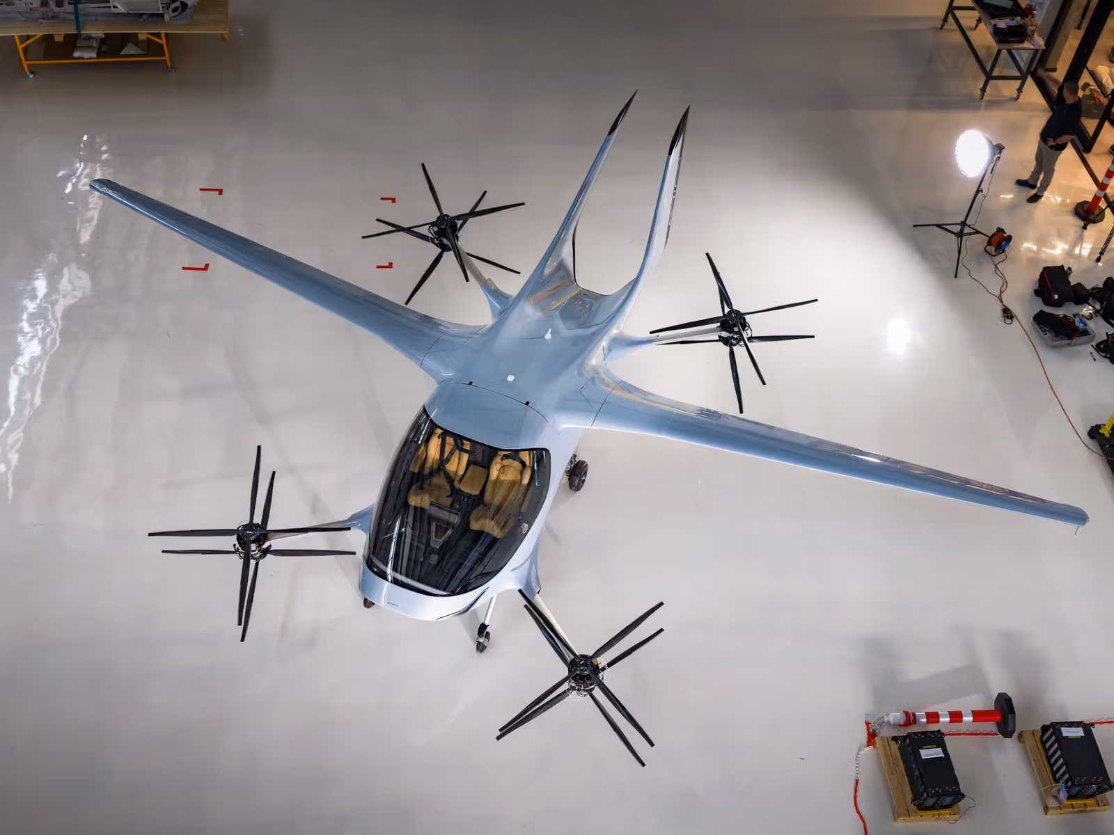Top view of a sleek electric aircraft with eight motors and a transparent cockpit inside a hangar.