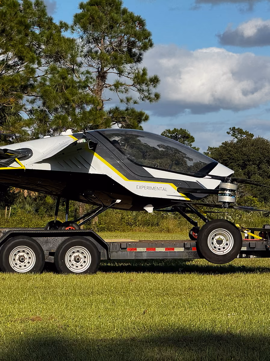 Experimental single-seat aircraft with wheels on trailer parked on grass with trees in background.