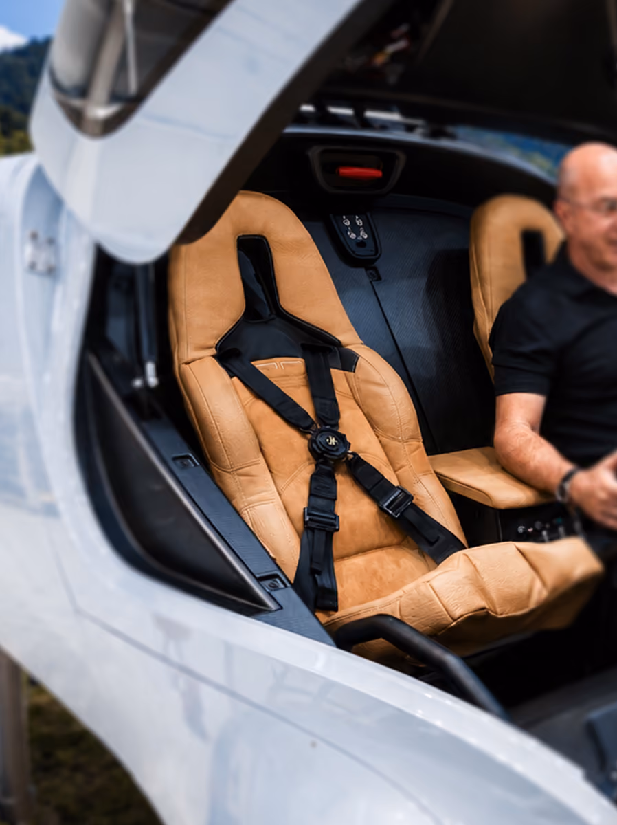 Tan leather racing seat with black harness inside a white vehicle cockpit.