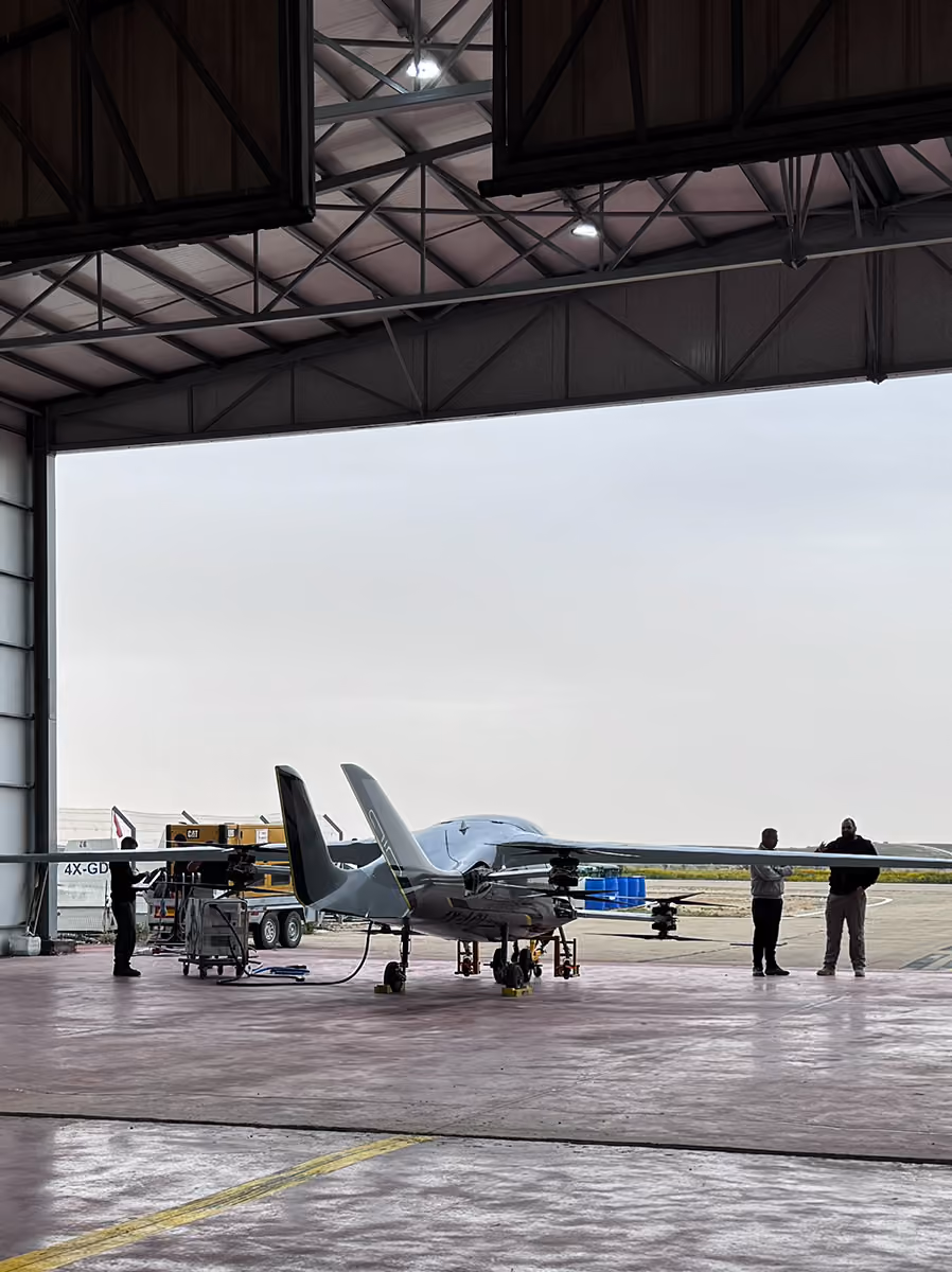 Small aircraft parked inside a hangar with three people standing near it on the tarmac.