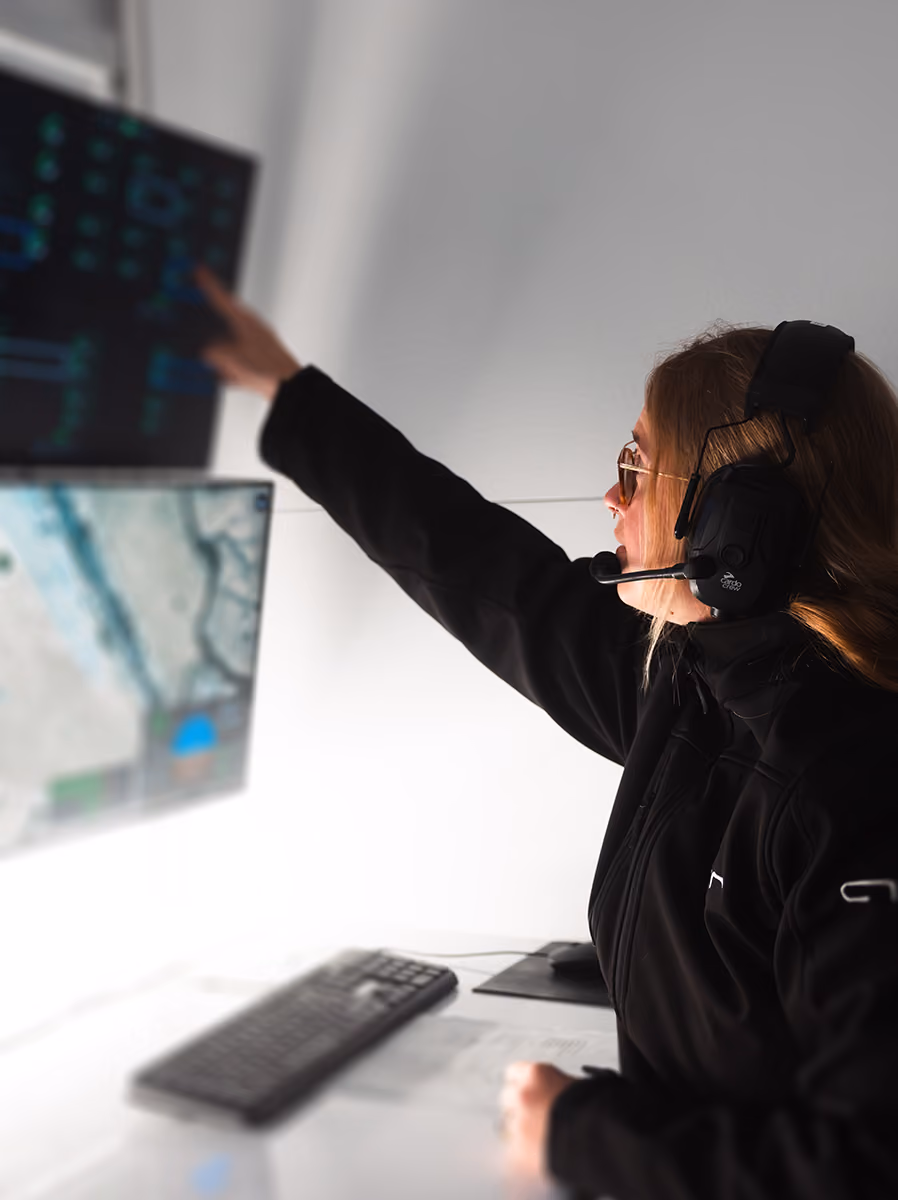 Woman wearing headset points at a screen with maps and data in a control room.