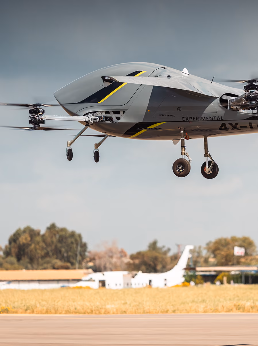Gray experimental VTOL aircraft hovering above runway with blurred background of trees and buildings.