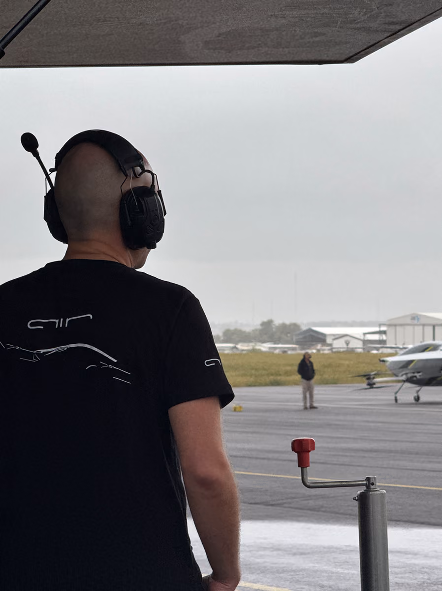 Man wearing headset and black shirt stands in a hangar, looking at a small plane on the runway.