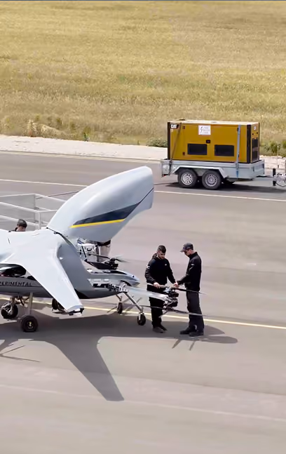 Two men inspect an experimental drone with open cockpit on an airstrip near a yellow equipment trailer.