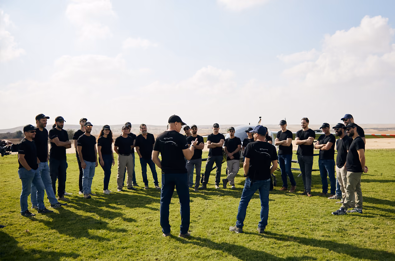 Group of people in black shirts and caps standing in a circle on grass near a small aircraft.