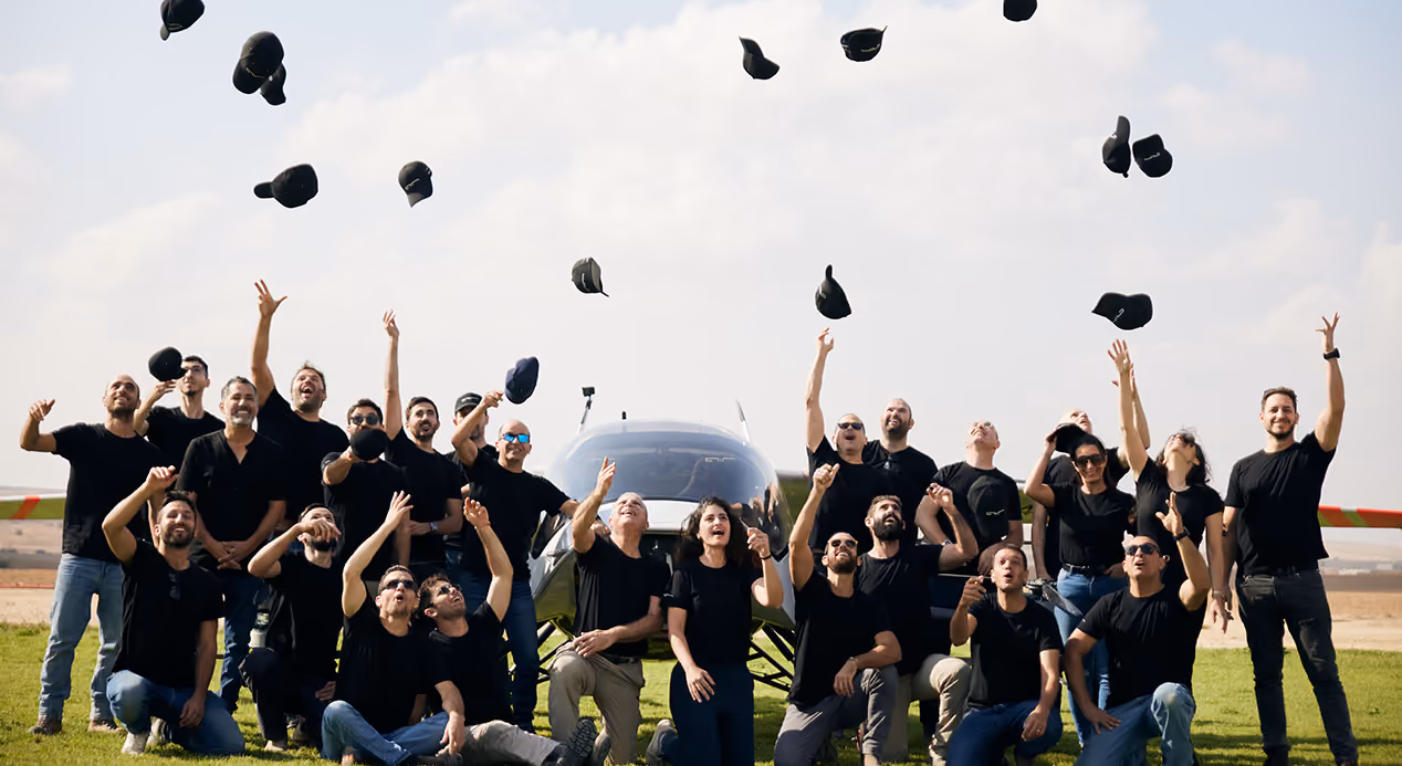 Group of people in black shirts tossing black caps in the air in front of a small aircraft on grass.