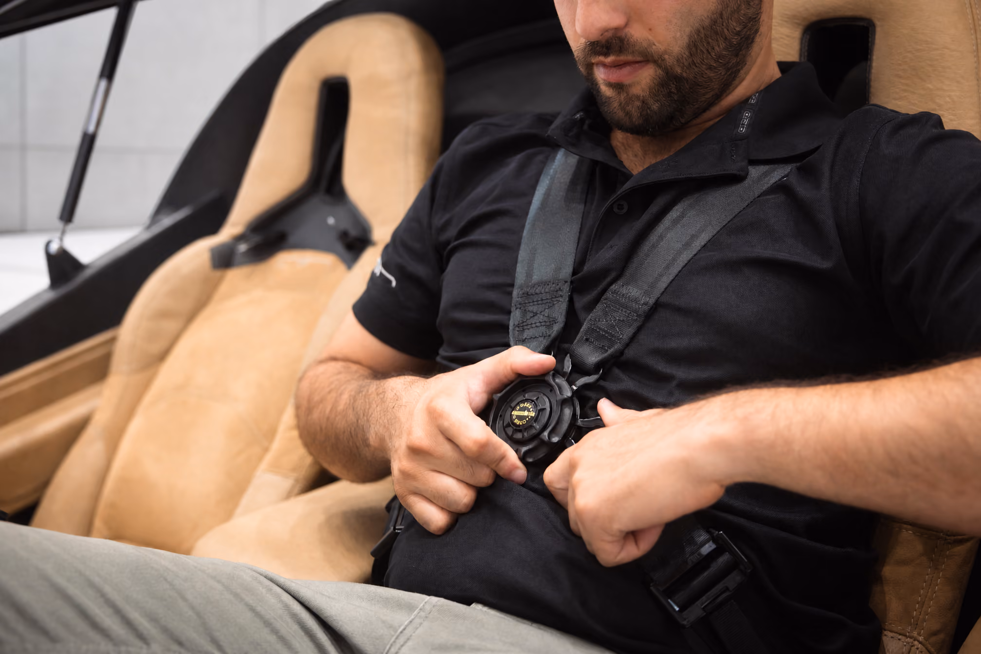Man fastening black four-point harness seatbelt while sitting in a tan vehicle seat.
