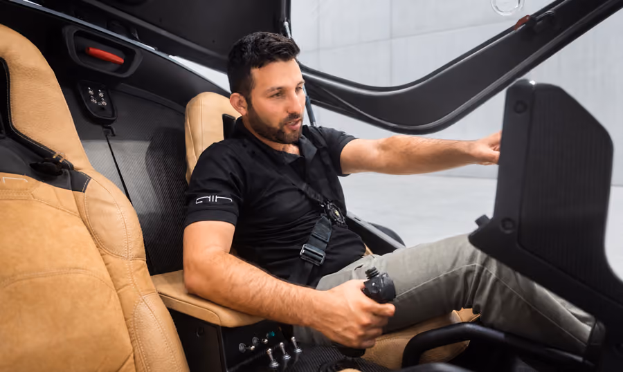 Man seated in a cockpit holding controls, wearing a black shirt and strapped in with a harness.