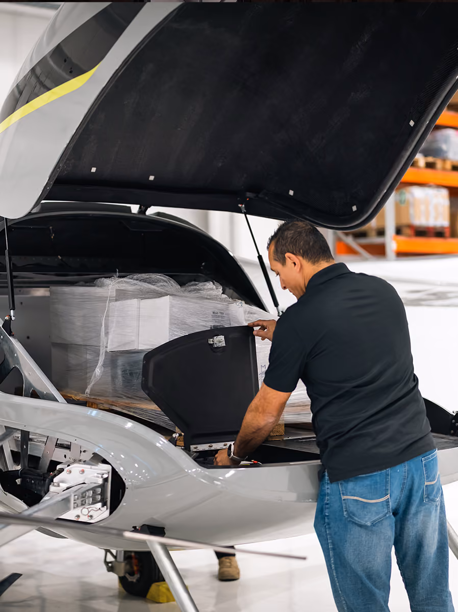 Man inspecting open front compartment of a light aircraft loaded with wrapped cargo inside a hangar.