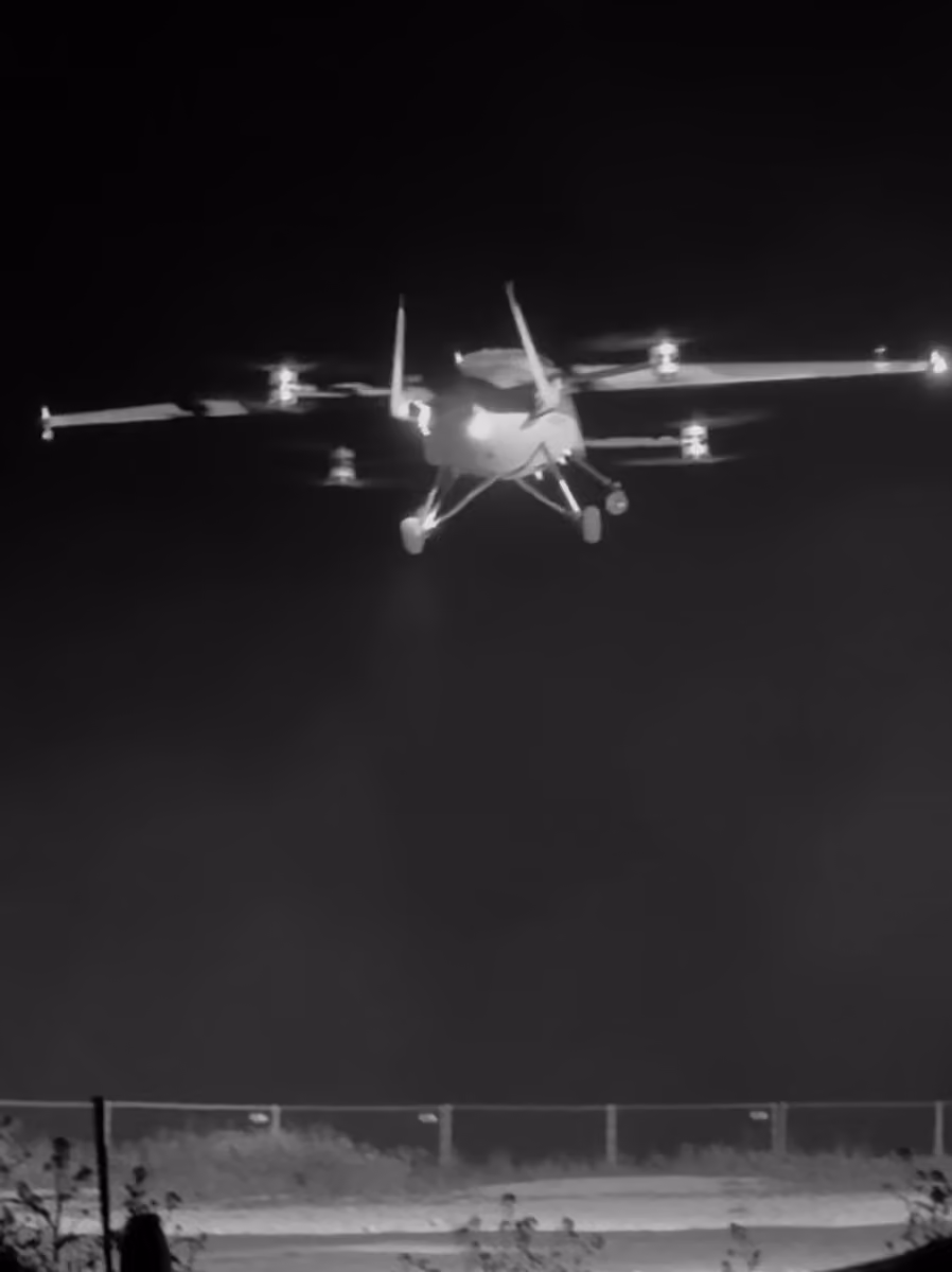 Black and white image of a drone or small aircraft flying at night over a fenced area.