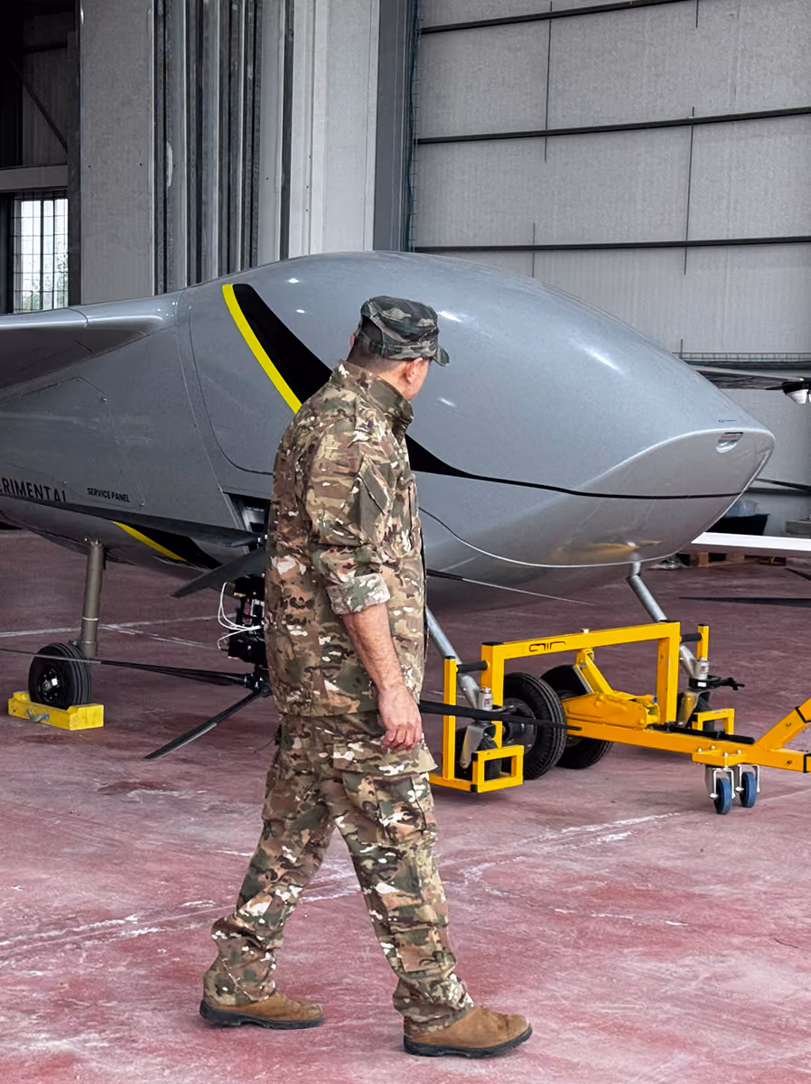Man in camouflage uniform near a gray experimental drone inside a hangar.