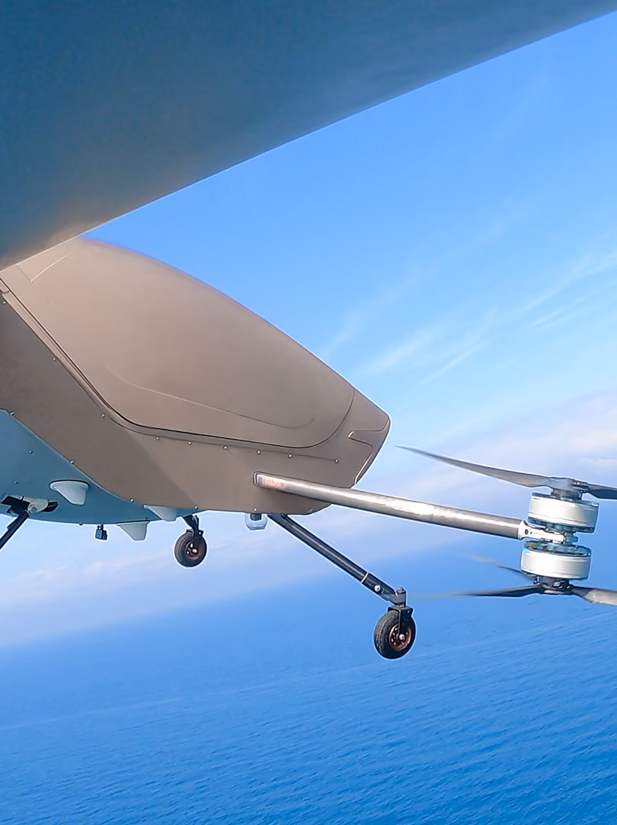 Close-up of a drone with rotors and landing wheels flying over a blue ocean under a clear sky.