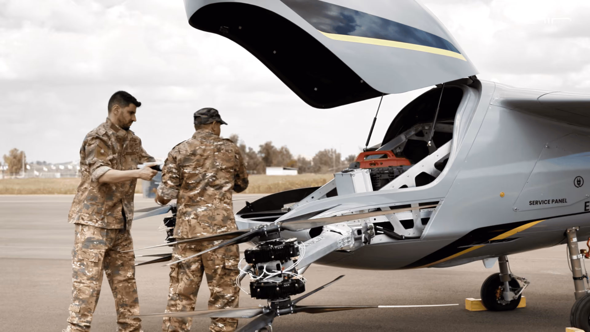 Two soldiers in camouflage uniforms work on parts of a helicopter on an airfield.