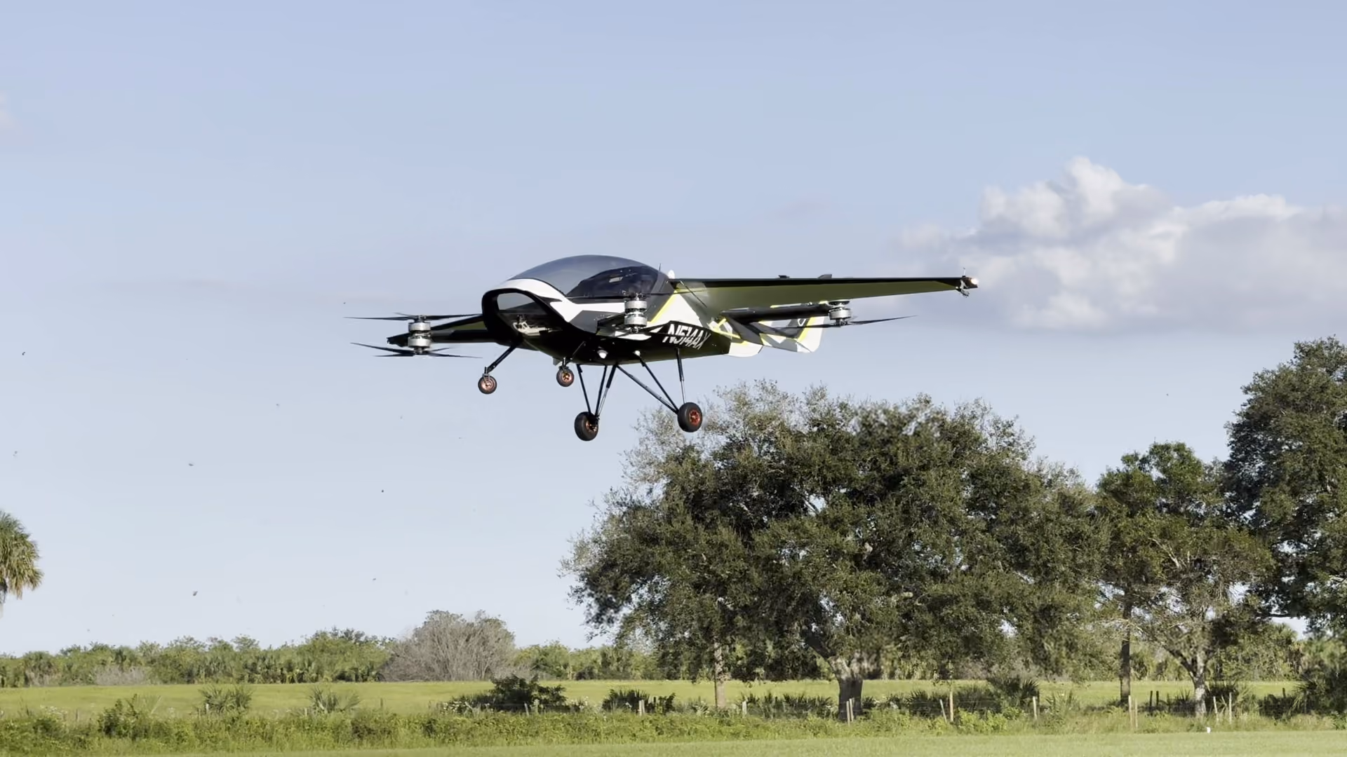 A black and white electric vertical takeoff and landing aircraft flying over grassy field with trees.