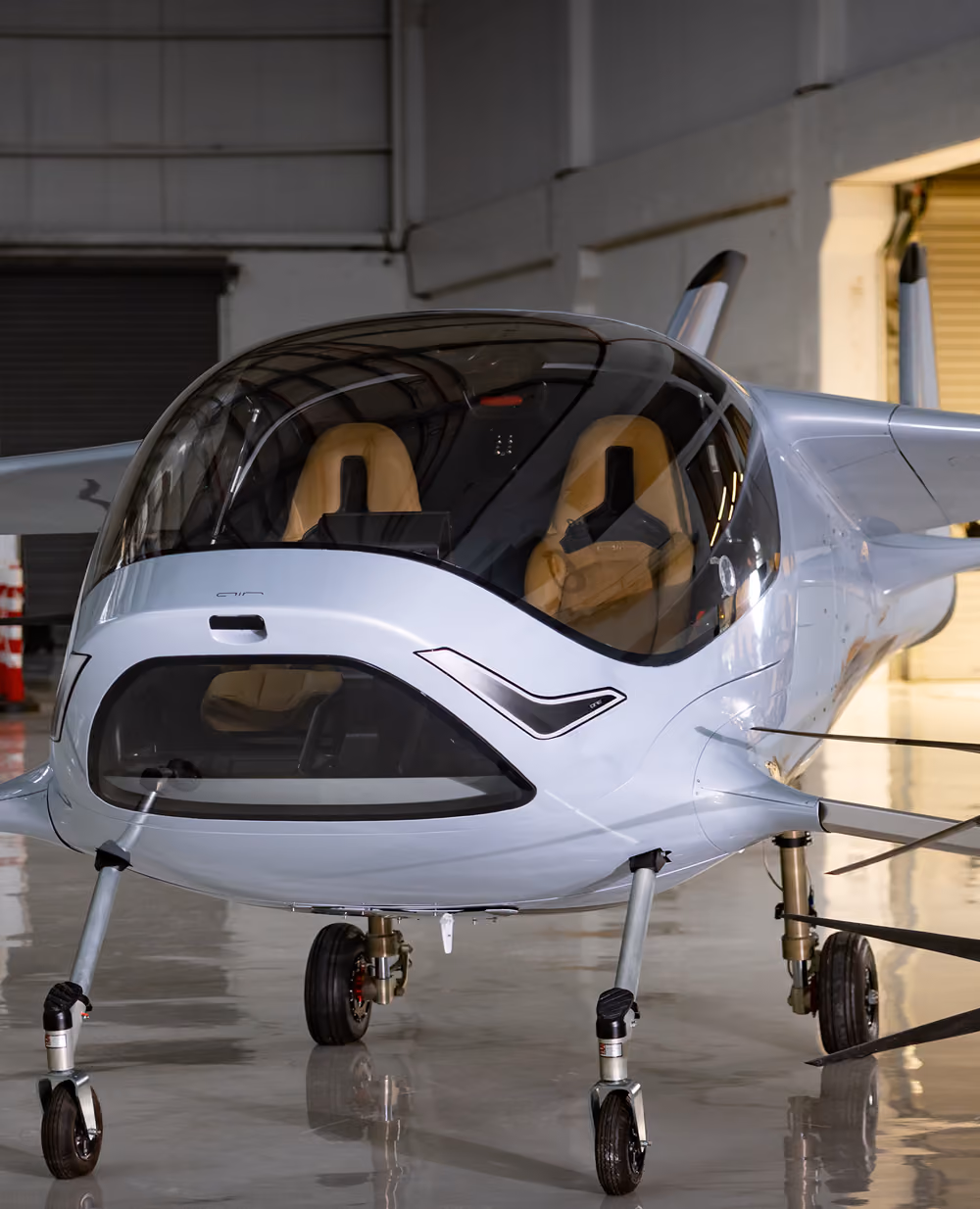 Front view of a sleek modern light aircraft with large windows and tan seats in a hangar.