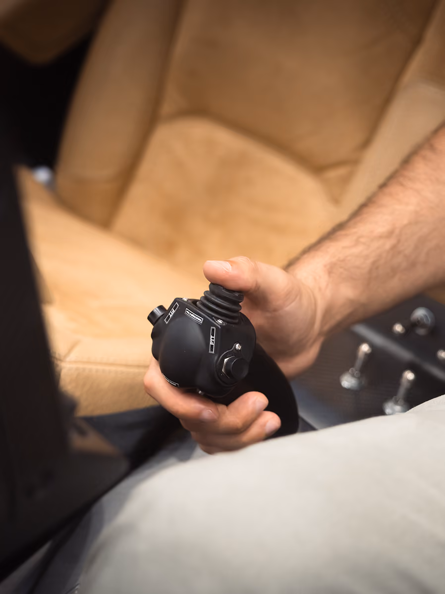 Close-up of a hand gripping a black joystick with buttons, mounted near beige seats and control panel.