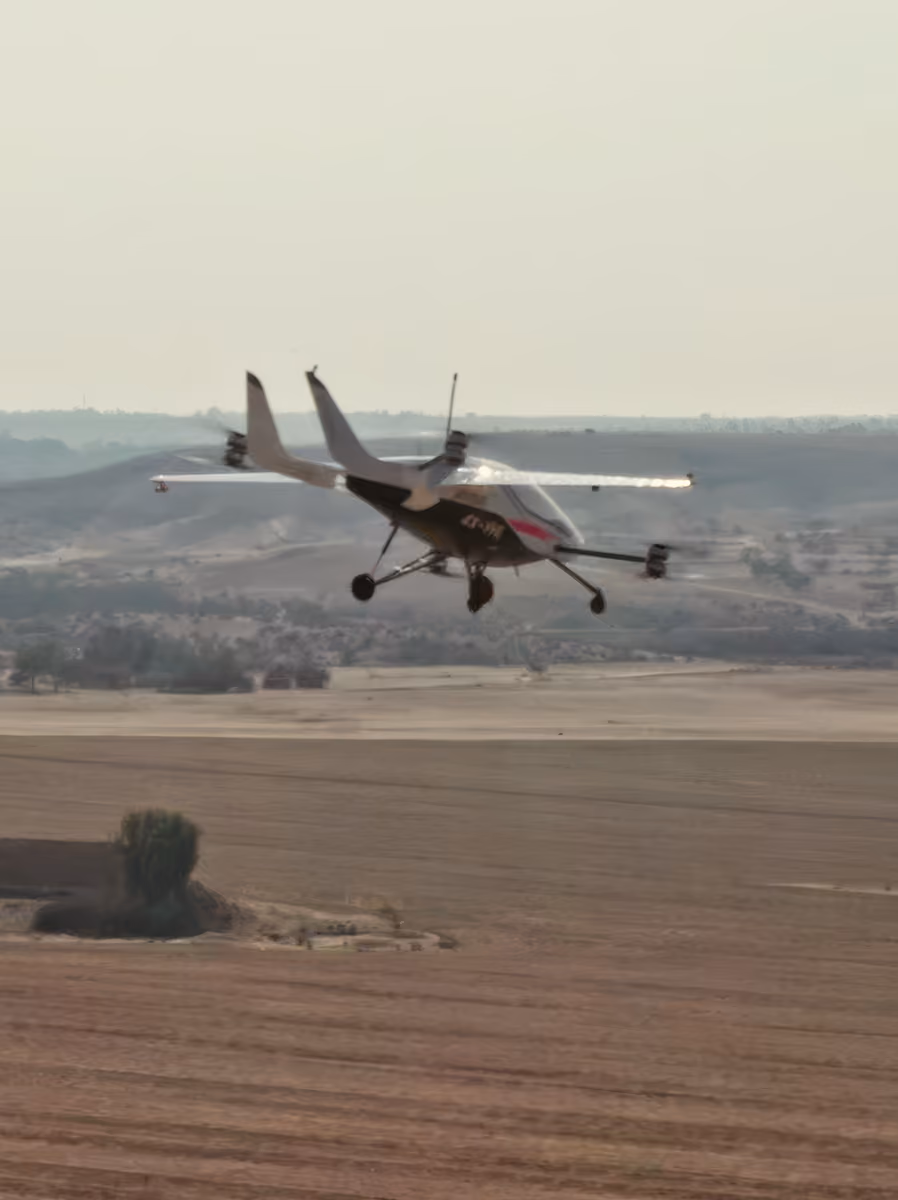 Small drone aircraft flying low over open farmland with hills in the background.