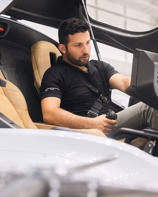Man with dark hair wearing a black polo shirt sitting in a car seat holding a joystick control.