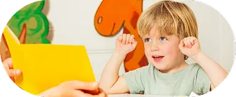 Young boy raising his hands while looking at an adult holding a yellow book.