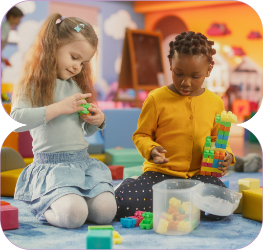 Two young girls sitting on a carpet playing with colorful building blocks in a bright classroom.