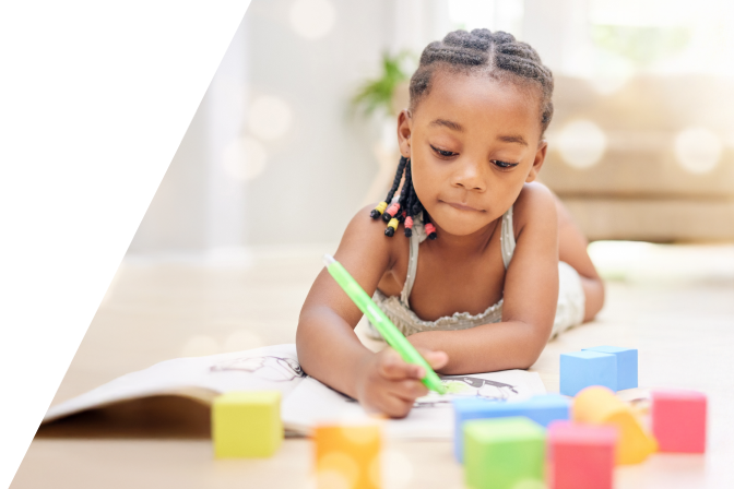 Young girl with braided hair coloring in a book while lying on the floor surrounded by colorful building blocks.