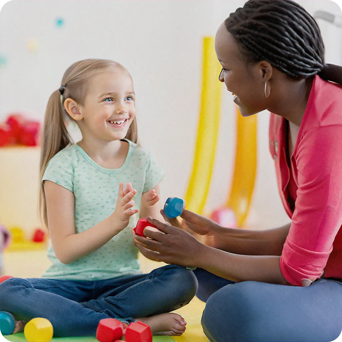 A smiling young girl with pigtails playing with colorful building blocks with an adult woman in a pink top.