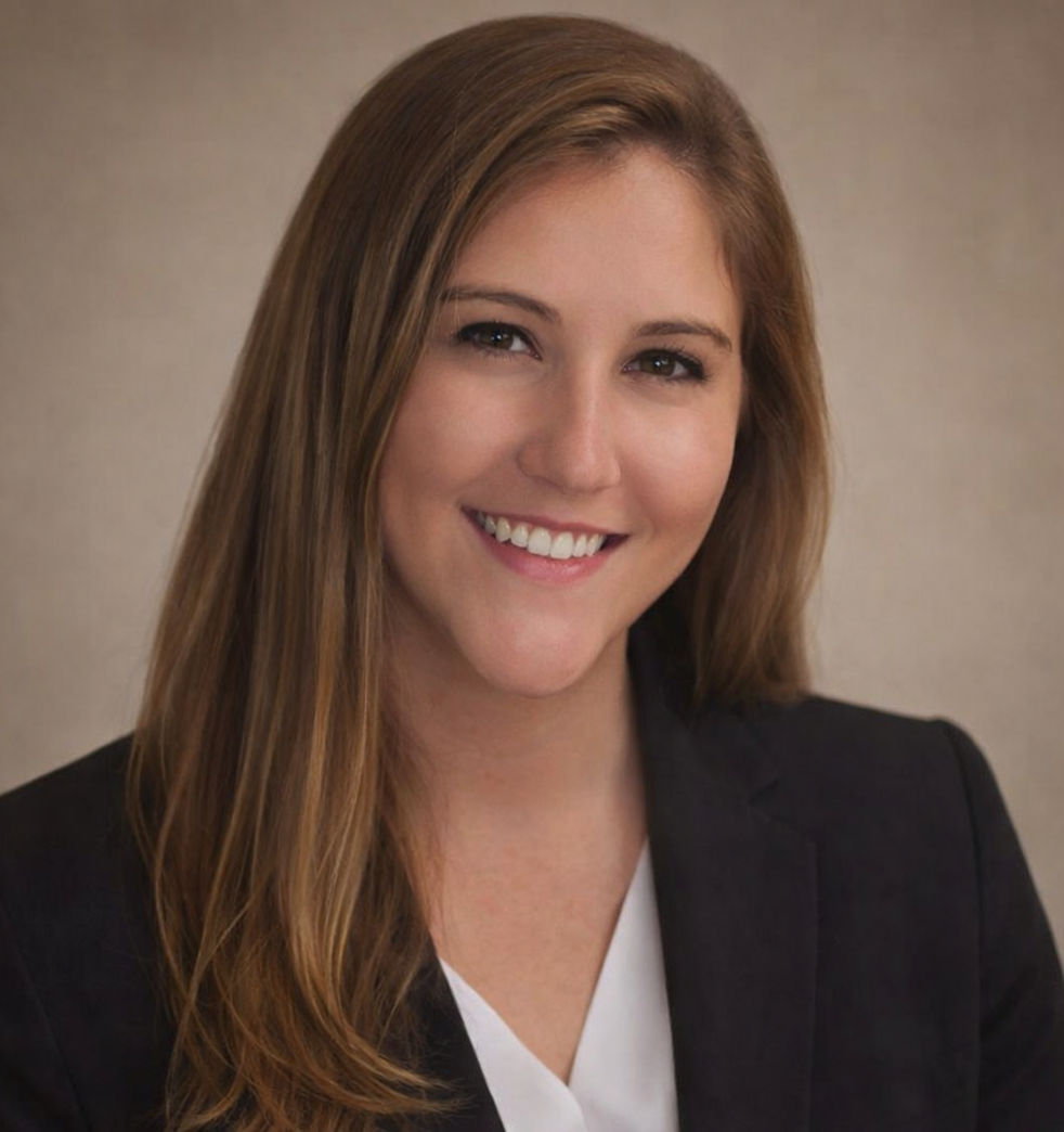 Smiling young woman with long brown hair wearing a black blazer and white blouse against a plain background.