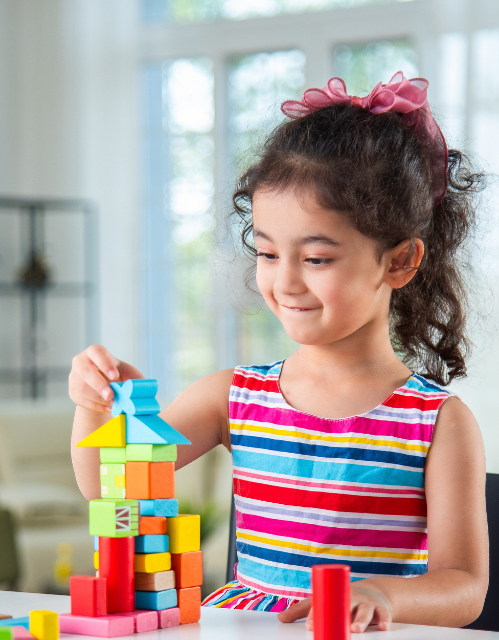 Young girl with a pink bow in her hair plays indoors, building a tower with colorful wooden blocks.