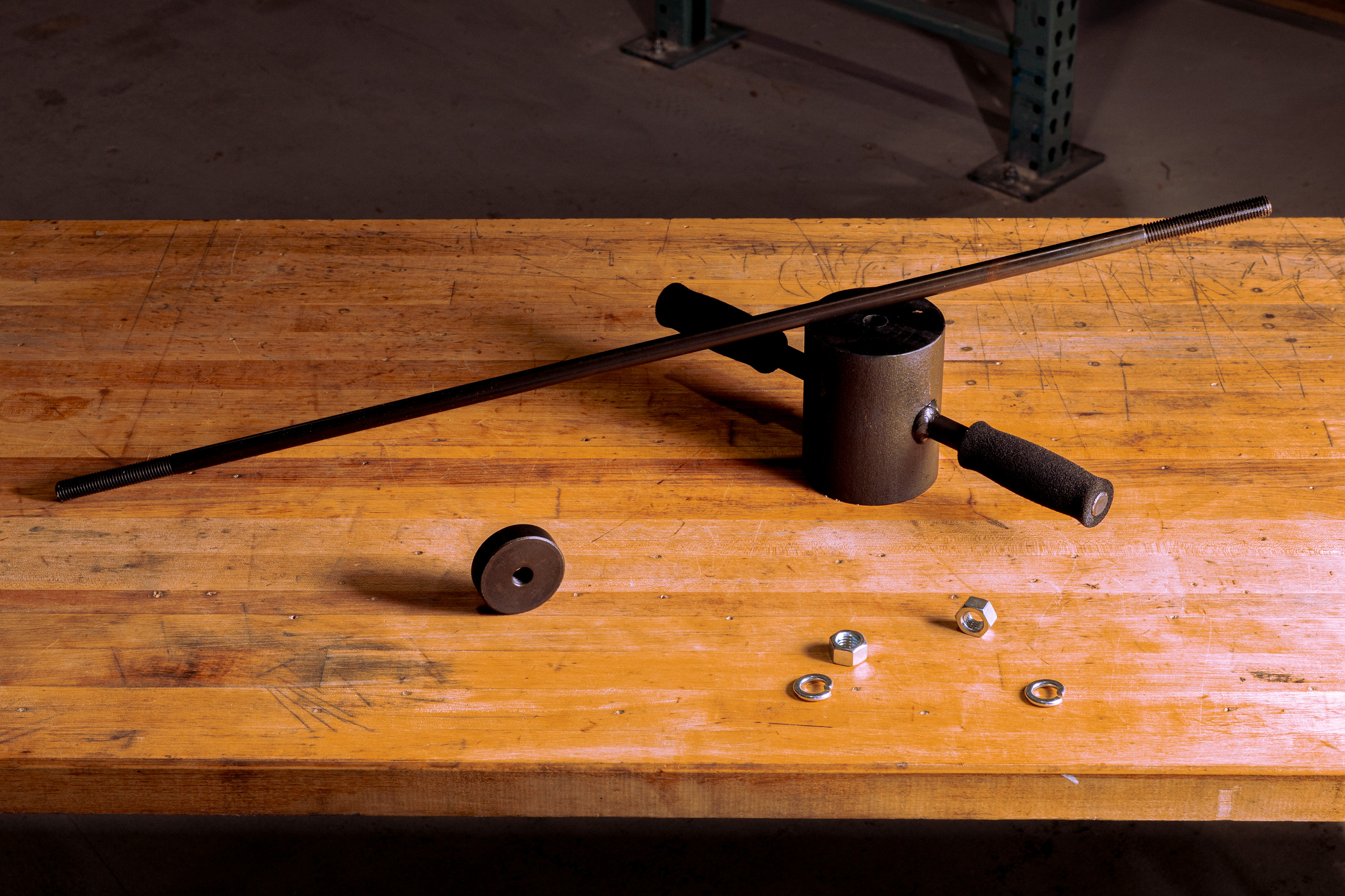 Metal threaded rod, cylindrical metal tool with a handle, two hex nuts, and two washers on a wooden workbench.
