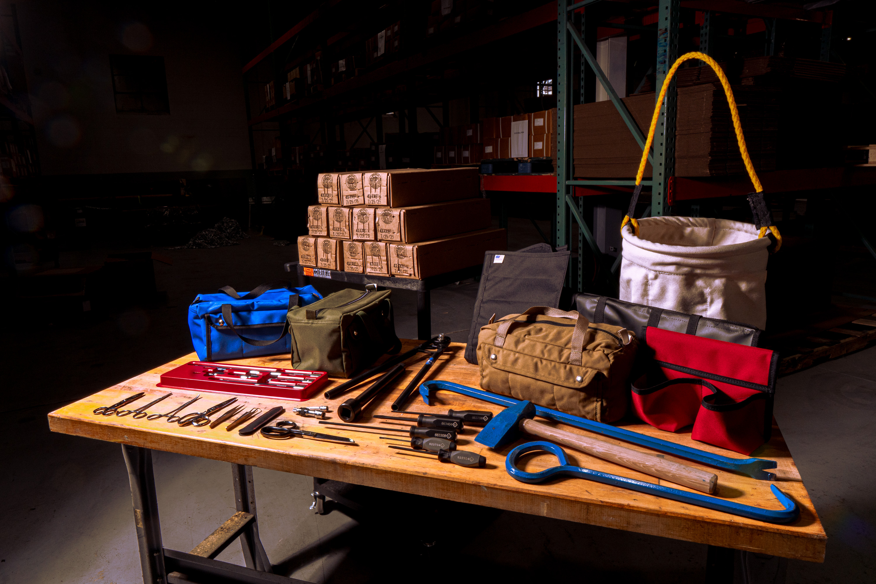 Wooden workbench with assorted hand tools, tool bags, and a white canvas bucket with yellow handles in a dimly lit warehouse.