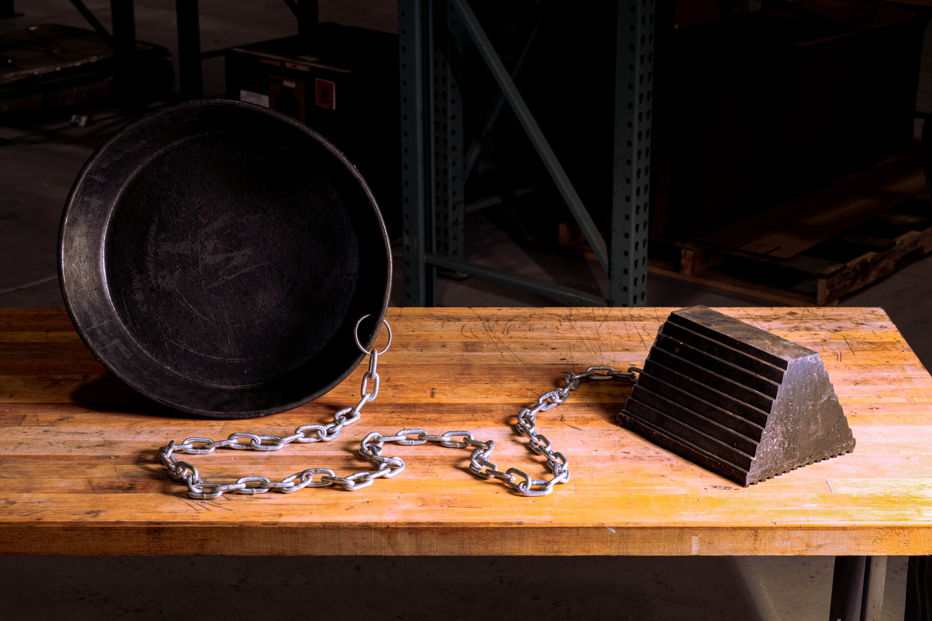 Metal bucket chained to a large metal wedge block on a wooden table in a workshop.