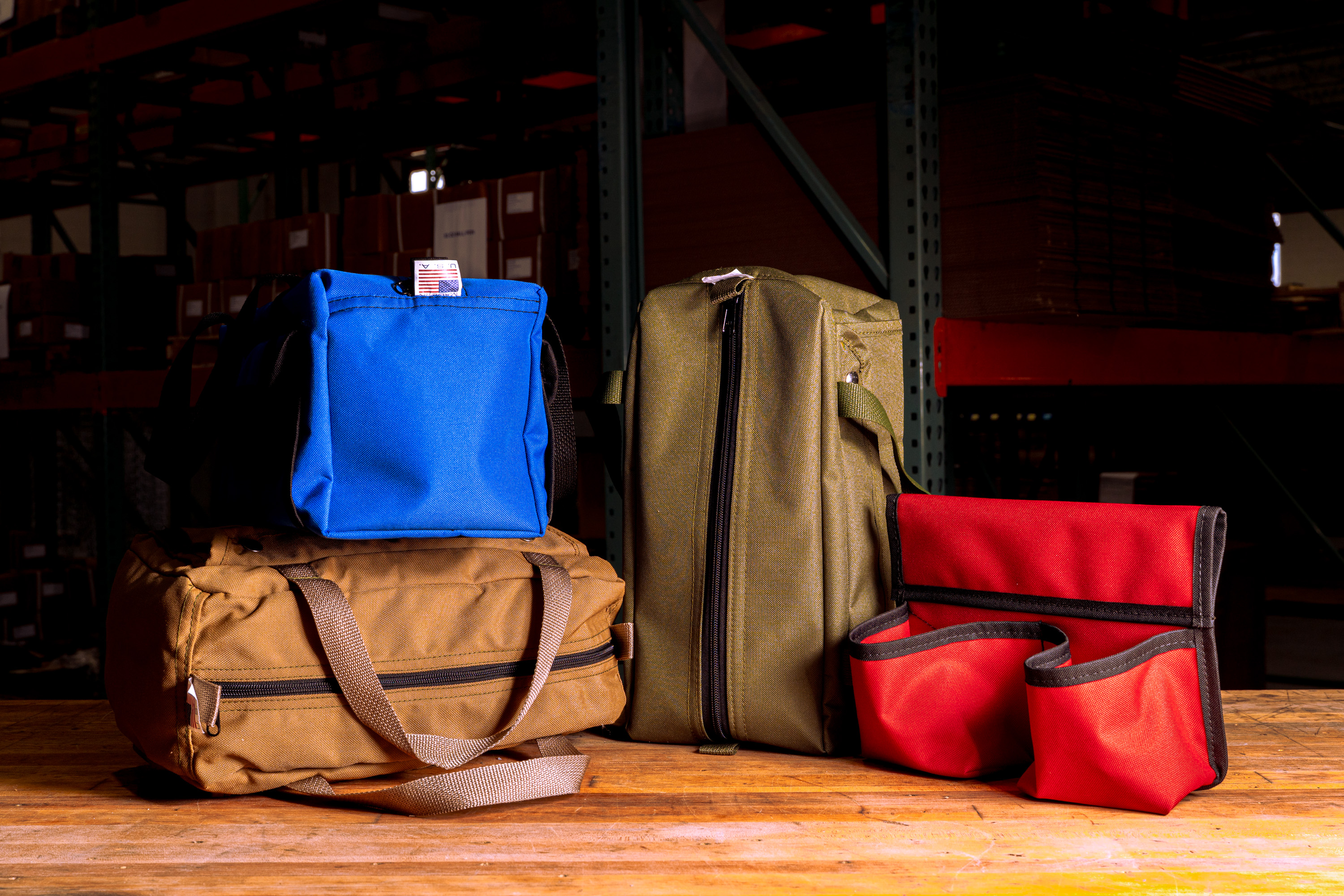 Four colorful fabric bags in blue, brown, olive, and red lined up on a wooden surface in a warehouse setting.