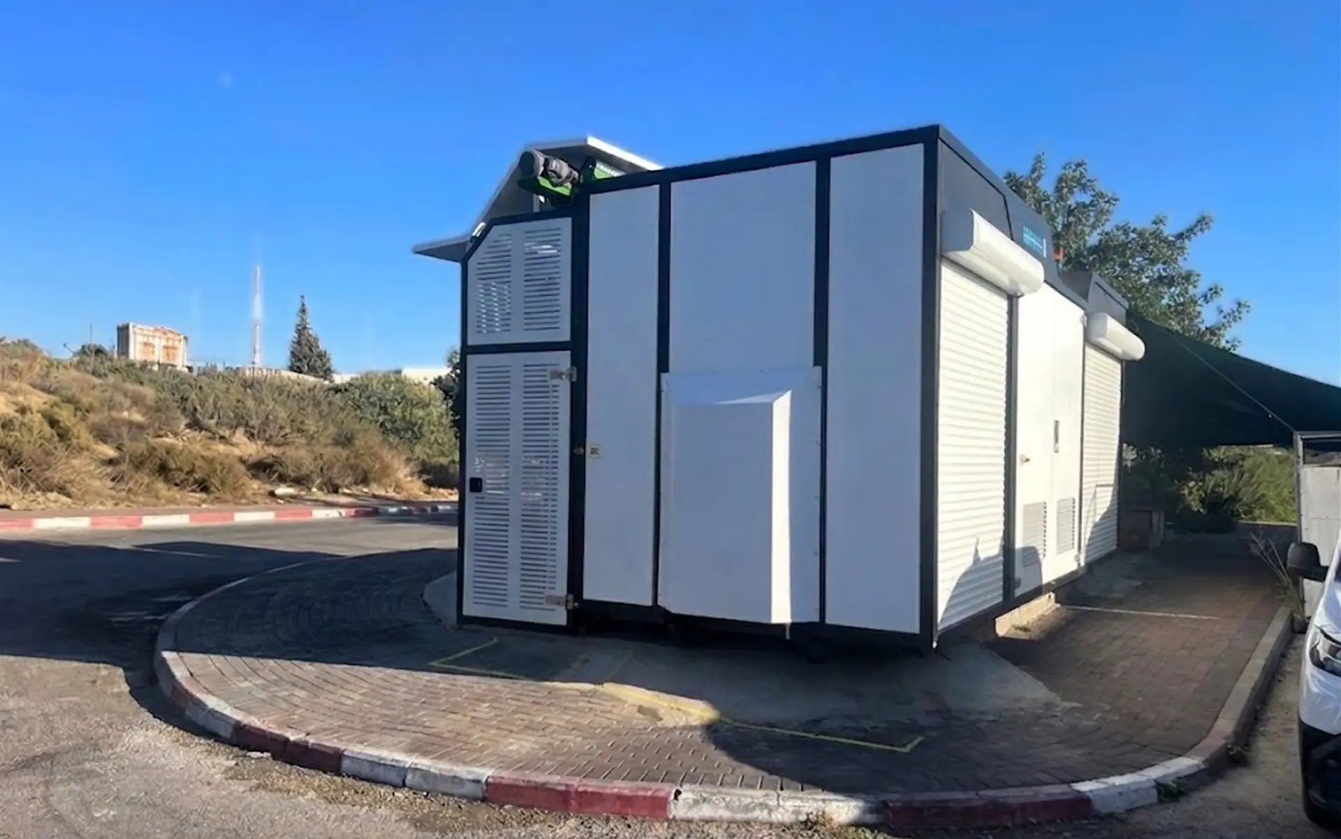 White and black modular utility building with vents and roller shutters on a paved corner with clear blue sky in the background.