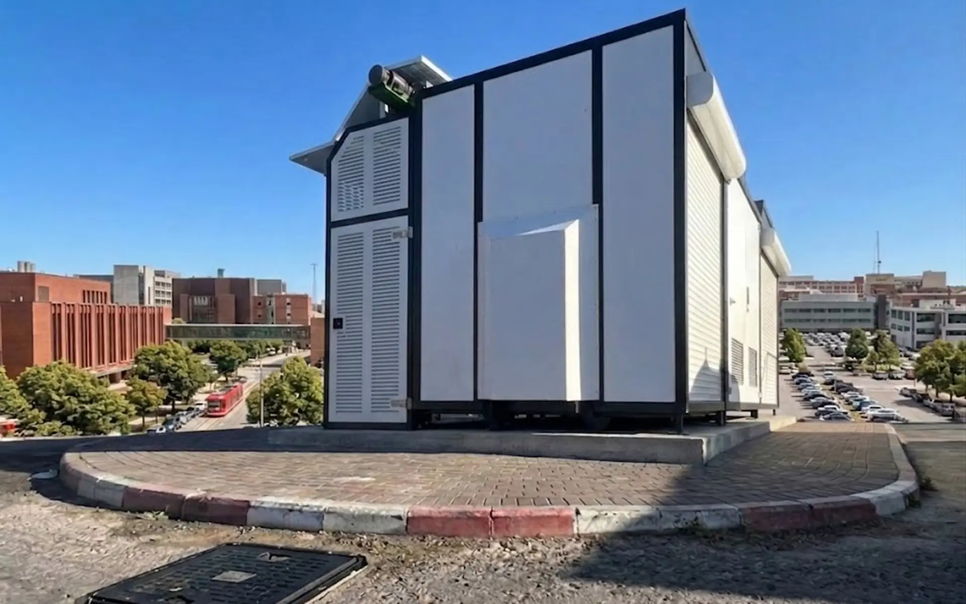 Large white industrial container or modular building on a raised platform with urban buildings and parked cars in the background under a clear blue sky.