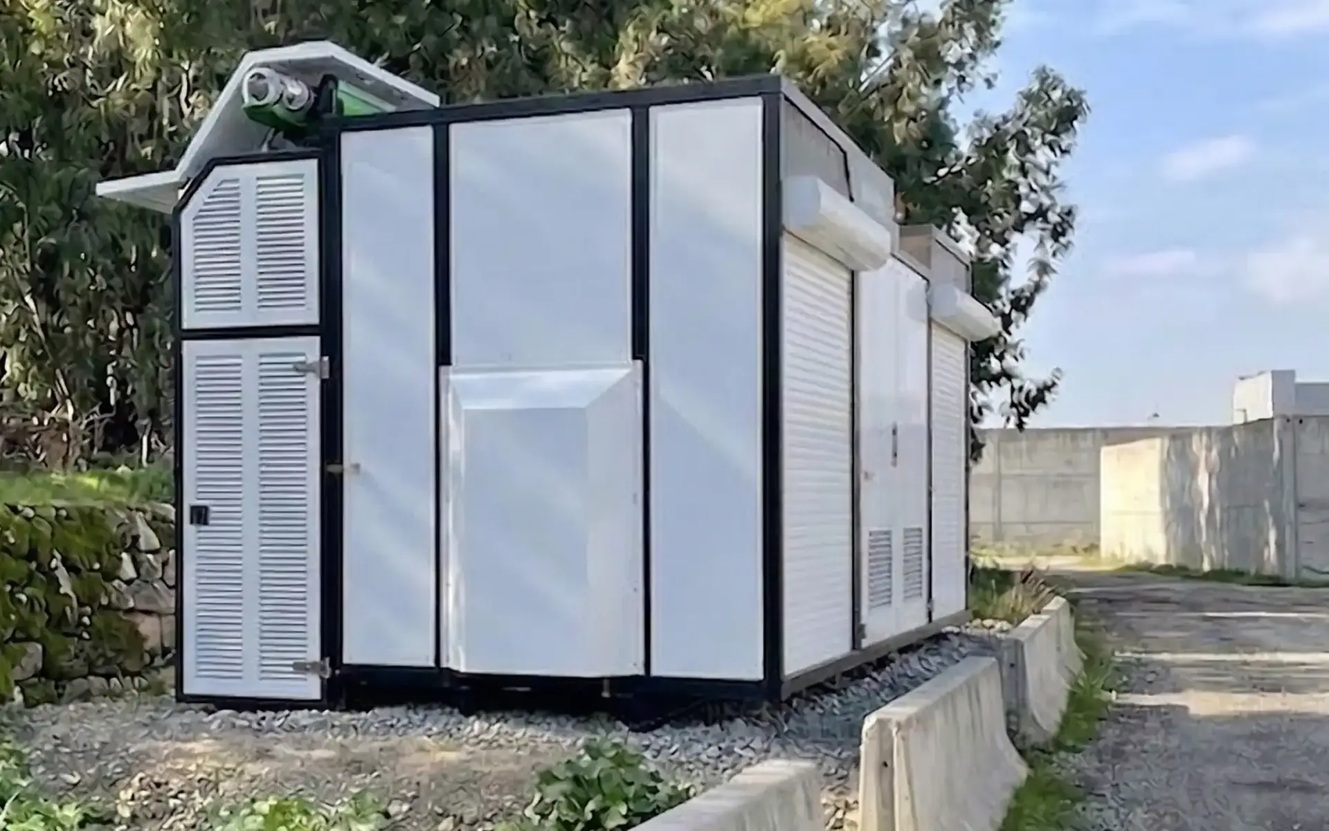 Rectangular white and black modular structure with vented doors and a small solar panel on top, located outdoors on a gravel base.