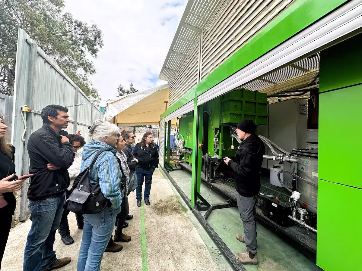 Group of people attentively listening to a man explaining equipment inside a large green industrial machine outdoors.