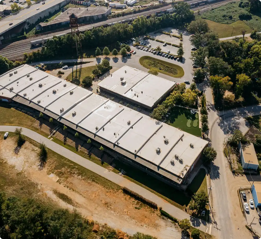 Aerial view of a commercial building complex with white flat roofs, surrounding parking lots, roads, and greenery.