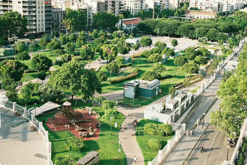 Aerial view of a city park with green trees, walking paths, a playground, and surrounding buildings.