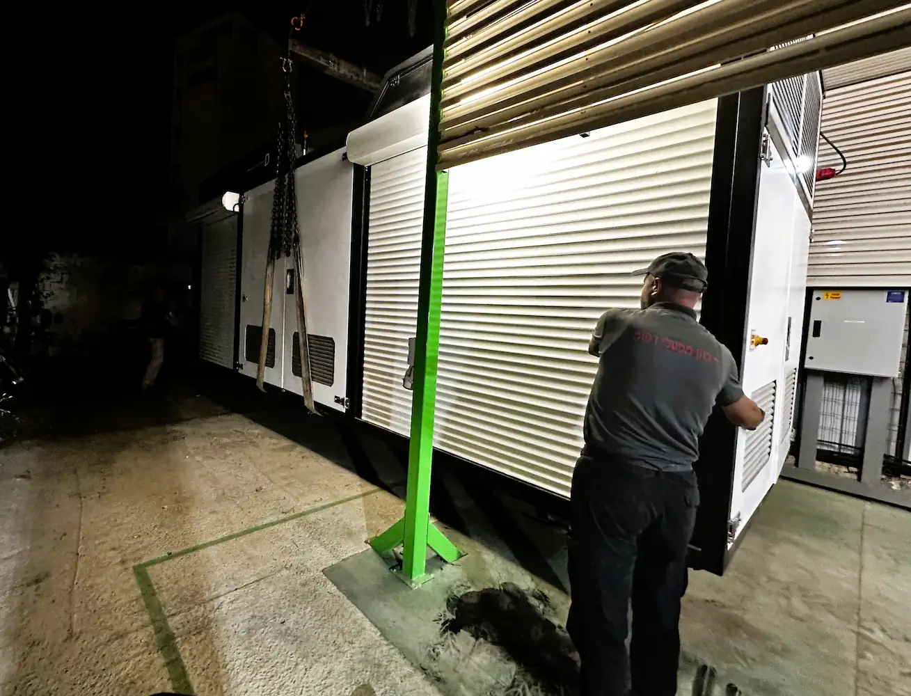 Man in dark clothing working on large white industrial equipment with rolling shutters at night.