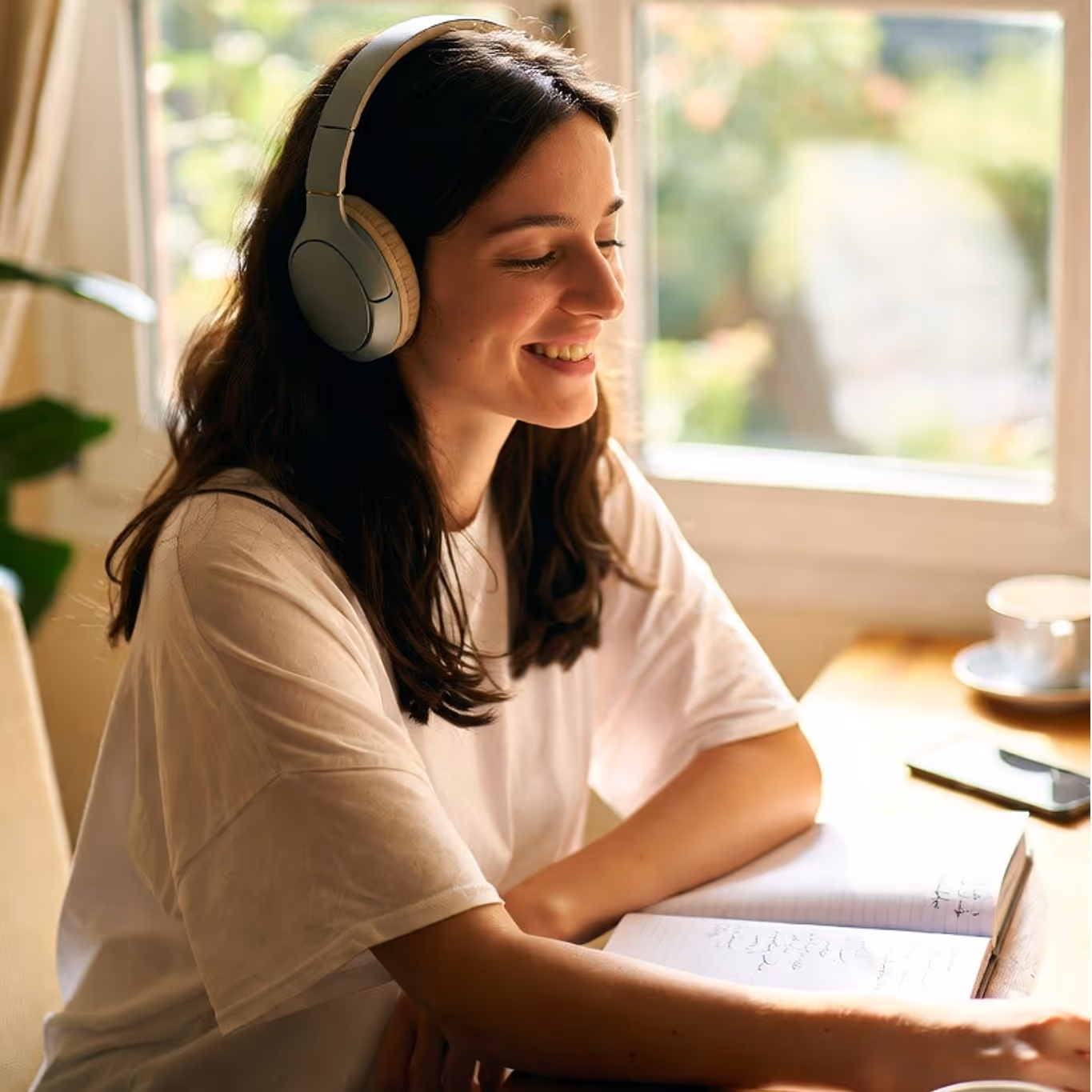 Jeune femme souriante portant un casque, assise à une table près d'une fenêtre avec un carnet ouvert.