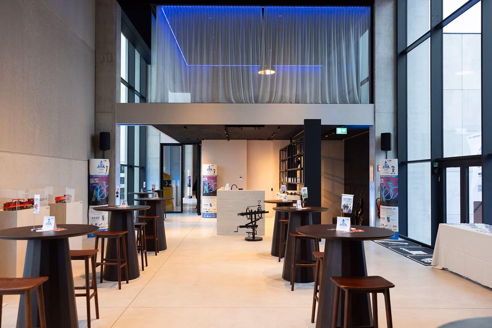 Modern lichte zaal met hoge ramen, hoge tafels en krukken, decor en heldere verlichting.