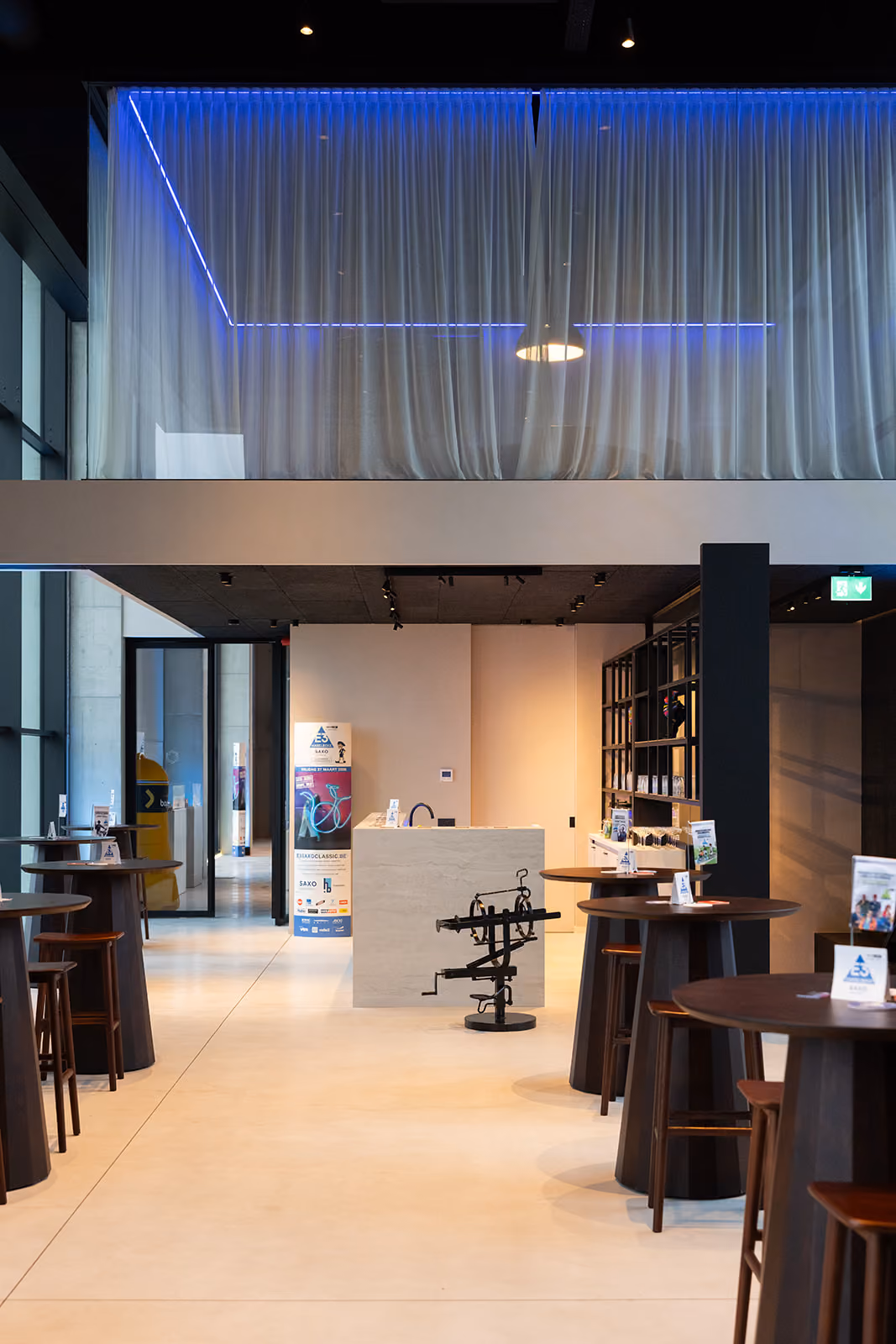 Modern binnenruimte met hoge tafels en stoelen, centrale balie en blauwe LED-verlichting onder vitrage.
