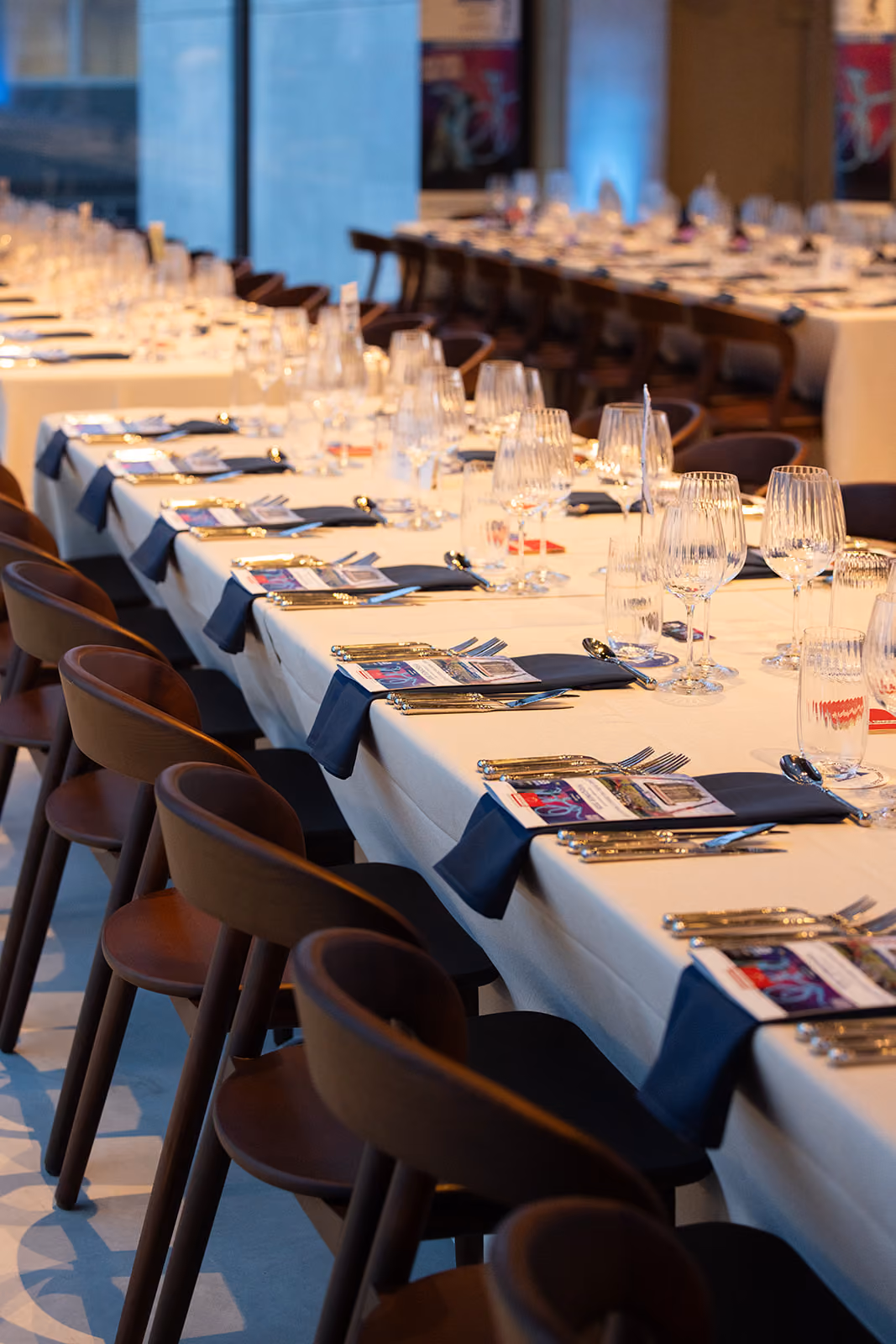 Lang gedekte tafels met glazen, bestek en donkerblauwe servetten in een sfeervolle eetruimte.